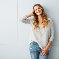 Beautiful mid-age woman standing by blue wall in office or home, smiling at camera. Casual clothes, caucasian. Long blond hair.