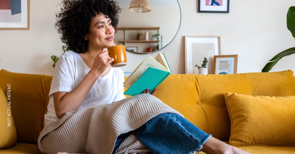 Young woman sits on yellow couch reaching a book while drinking from a mug. (Model)