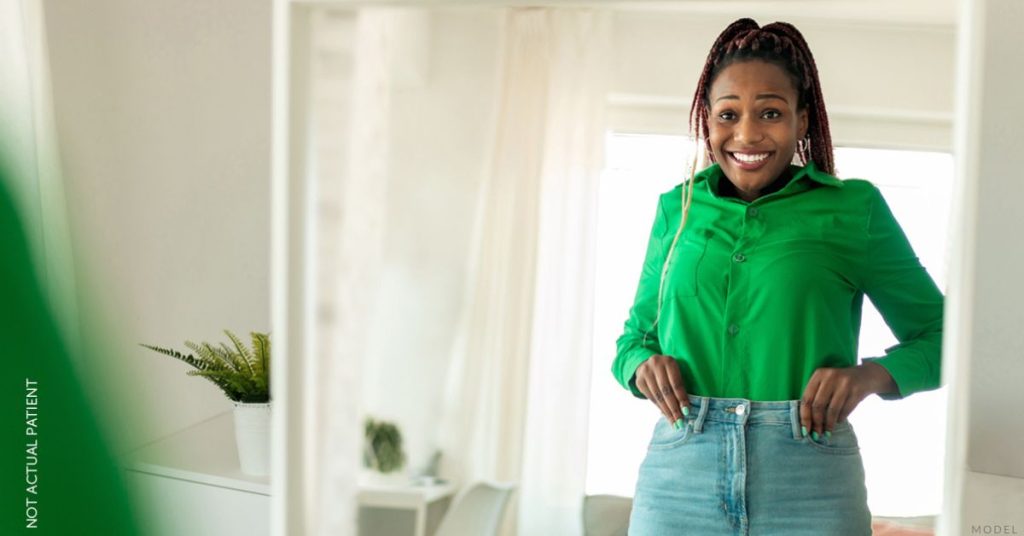 A woman adjusts her jeans, looking at herself in the mirror with a smile (MODEL).