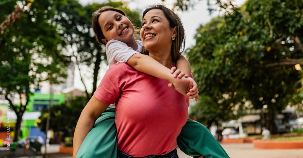 Smiling woman giving a piggyback ride to a young girl outdoors, representing life after weight loss skin tightening (not actual patients)